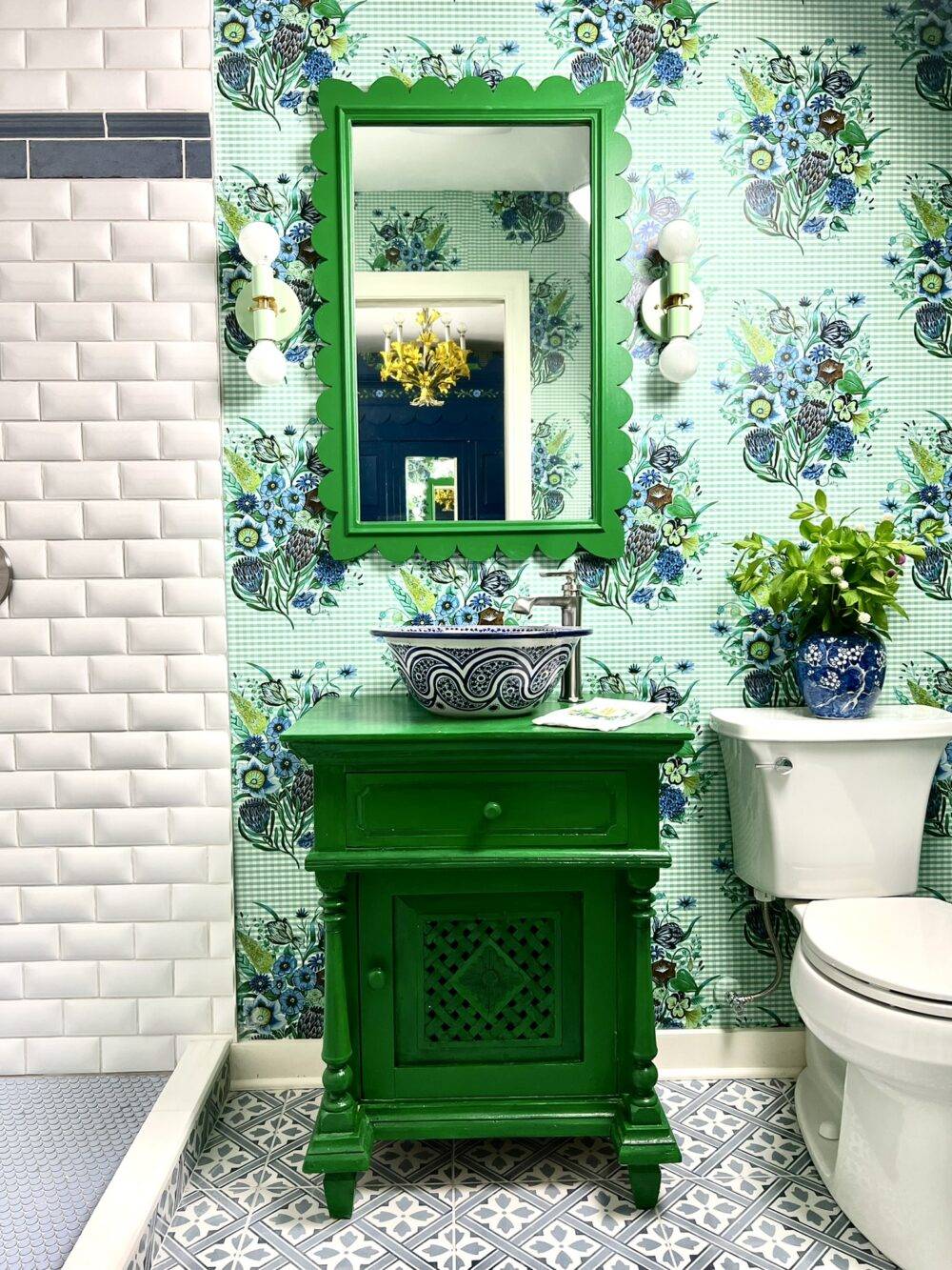 Blue and green bathroom with floral wallpaper, green sink and mirror and patterned floor tile. 