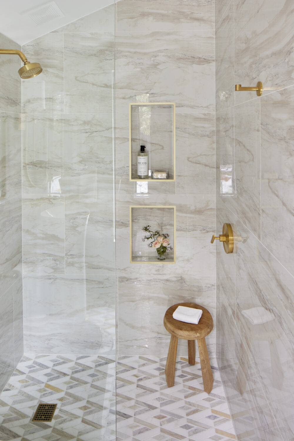 Shower with beige marble-look wall tile and gold trimmed shower niches and mosaic marble floor. 