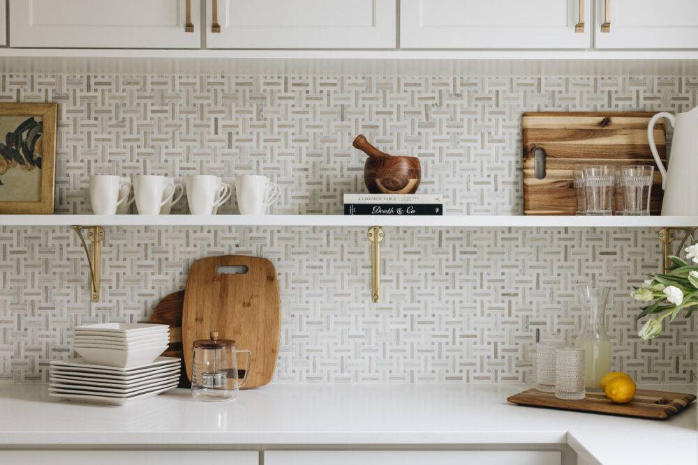 Kitchen counter and shelf with cups, cutting board and other cooking elements before white and beige marble basketweave tile.